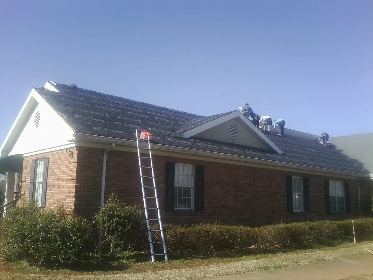 Crew tearing shingles off a brick ranch roof with a ladder set — Metal Roof Repair work by our crew in North Zulch