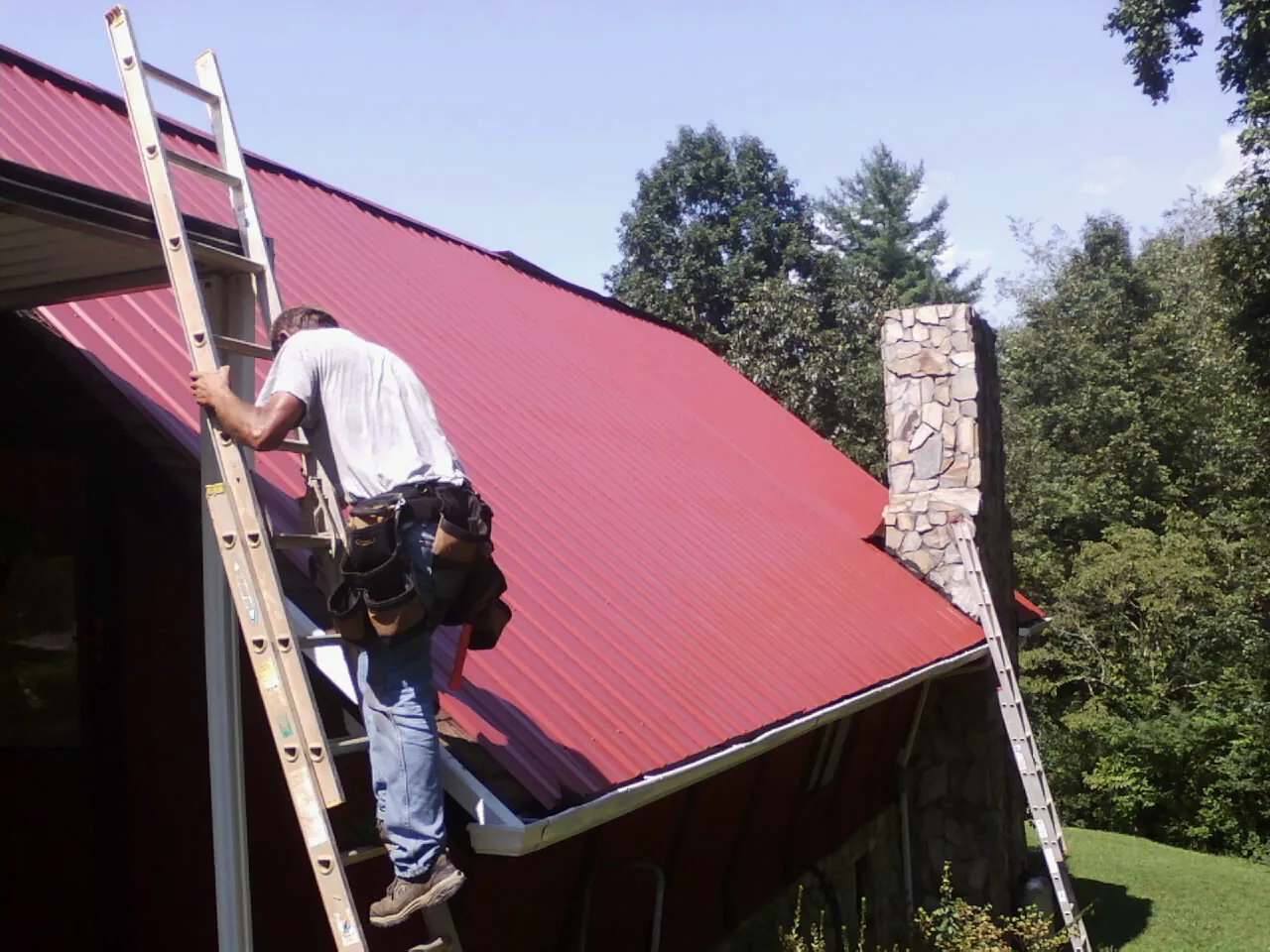 Roofer on a ladder beside a red metal roof and stone chimney — Soffit & Fascia Repair by Roofing Brothers in North Zulch